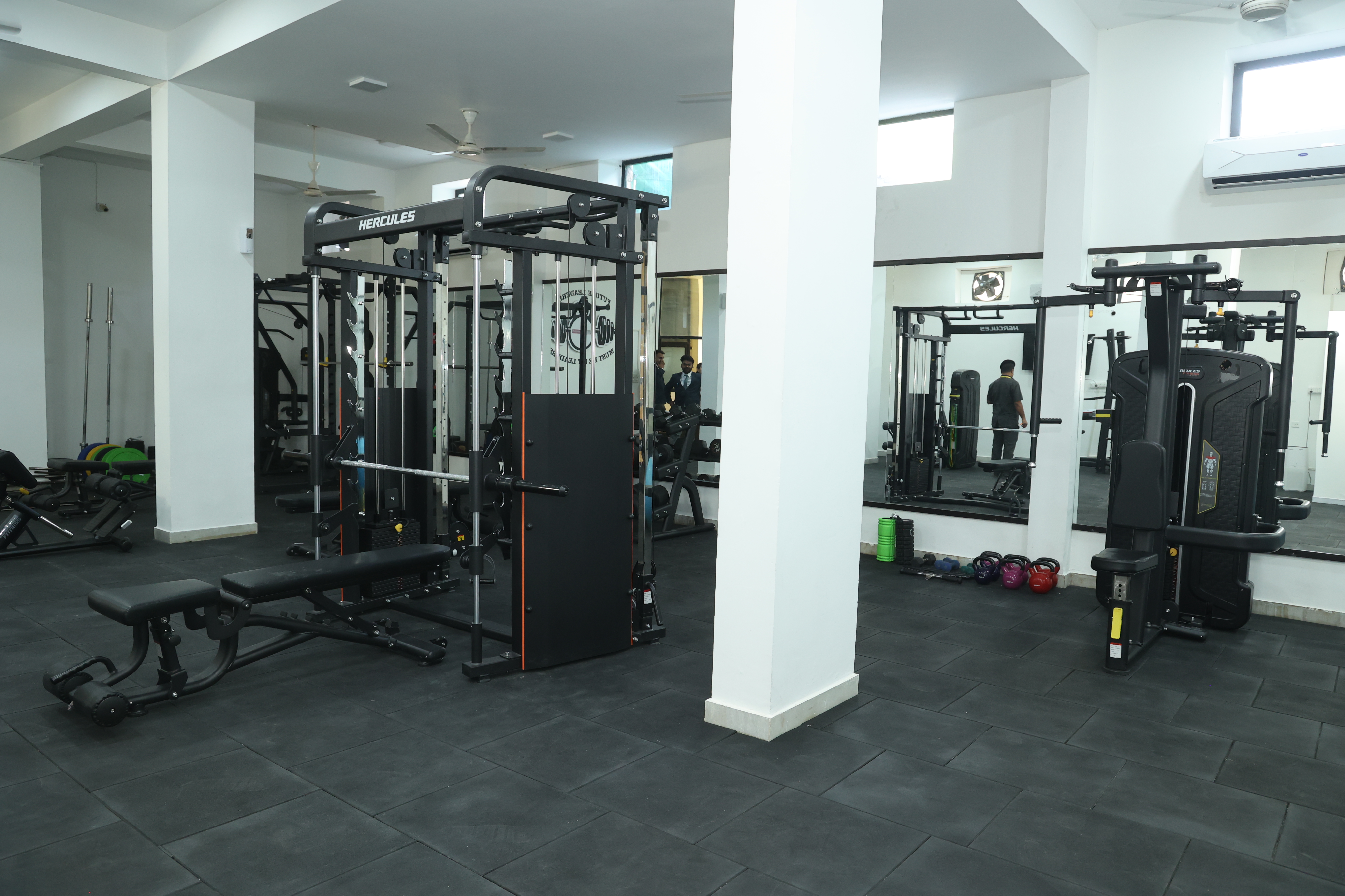 A well-equipped gym of IIHMR University Jaipur with various exercise machines, weights, and a blue exercise ball on a red carpeted floor. Fitness posters adorn the walls